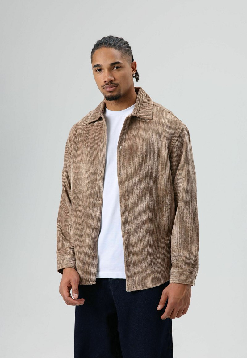 Brown corduroy shirt with a collared neckline, button closure, and long sleeves, layered over a white T-shirt. Textured surface with vertical ribs.