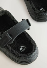 Schwarze, mit Fleece gefütterte Clogs mit verstellbaren Riemen und strukturierten Sohlen, die ein Innenlabel mit einem Berglogo und Text zeigen.