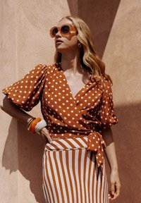 Haut enveloppant à pois marron avec des manches bouffantes, associé à une jupe rayée. Les accessoires comprennent des lunettes de soleil orange et des bracelets.