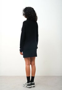 Zwarte gebreide trui met een col, gecombineerd met een zwarte rok. Model draagt zwarte sokken en zilverkleurige sportschoenen. Eenvoudig, ontspannen ontwerp.