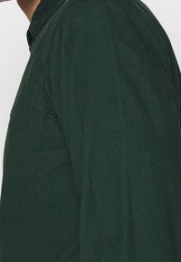 Tmavě zelená kordurová košile s texturovaným povrchem, která má klasický límec a boční pohled ukazující švy a detaily střihu.