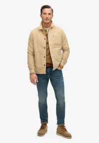 Beige katoenen jas met knoopsluiting, gedragen over een bruine gebreide trui. Gecombineerd met blauwe jeans en beige veterschoenen.