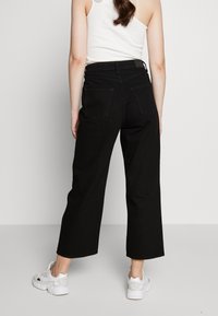 Schwarze weit geschnittene Jeanshosen aus Denim mit hoher Taille, zwei Gesäßtaschen und sauberem Finish. Kombiniert mit weißen Sneakern.