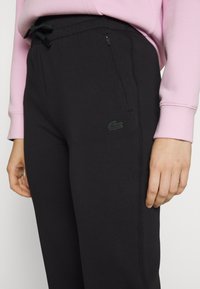 Svarte joggebukser i mykt stoff med elastisk midjebånd, med grønn logo-broderi og en sidelomme med glidelås.