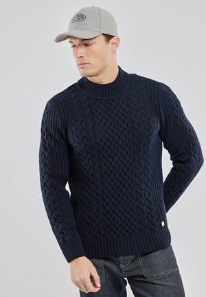 Homme portant un pull torsadé bleu marine et une casquette gris clair, regardant vers le bas avec les mains derrière le dos sur un fond uni.