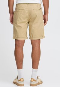 Beige Baumwollshorts mit umgeschlagenen Säumen, zwei Gesäßtaschen und einem Etikett am Bund. Getragen mit weißen Socken und beigen Sneakers.