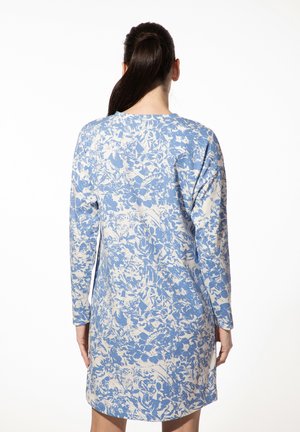 Vrouw met donker haar in een paardenstaart, gekleed in een abstracte bloemenjurk met lange mouwen in blauw en wit, gezien vanuit de achterkant tegen een witte achtergrond.