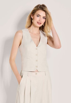 Frau trägt beige ärmellose, geknöpfte Weste und dazu passende, hoch taillierte Hosen mit Plisseefalten, posiert mit einer Hand in der Tasche und der anderen, die ihr Haar berührt.