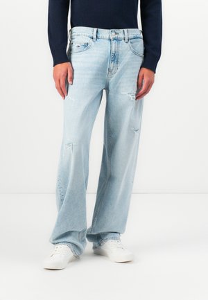 Persoon in lichtblauwe ripped wide-leg jeans, witte sneakers en een lange mouwen marineblauw top staande tegen een witte achtergrond.