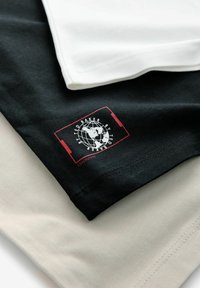 Tres camisetas apiladas: negra, blanca y beige. Tela de algodón suave. La camiseta negra presenta un logo rojo y blanco con un diseño de globo.