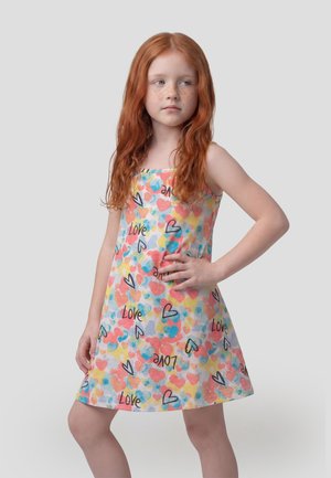 Niña pelirroja con pecas, vestida con un vestido sin mangas estampado con corazones multicolores y la palabra "Amor", de pie con una mano en la cadera.