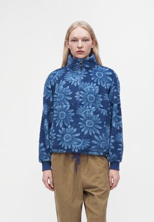Jeune femme aux longs cheveux blonds portant un pull polaire bleu à motif floral et un pantalon beige, debout devant un fond blanc uni.