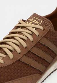 Καφέ αθλητικό παπούτσι με υφασμένη υφή, καφέ σουέντ λεπτομέρειες, ελαφρώς ανοιχτό καφέ κορδόνια και χρυσό λογότυπο. Διαθέτει τρεις χαρακτηριστικές ρίγες.