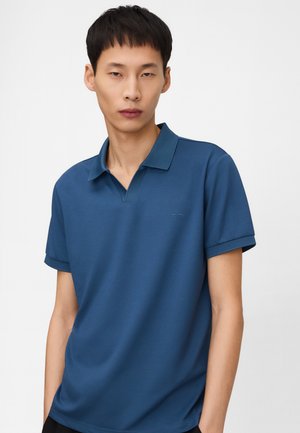 Junger Mann mit kurzem schwarzen Haar, der ein einfarbiges blaues Poloshirt mit kurzen Ärmeln vor weißem Hintergrund trägt.