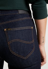 Jeans de mezclilla azul oscuro con una etiqueta de cuero negro, costuras naranjas y un diseño de bolsillo distintivo. Estética minimalista sin adornos.