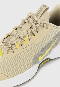 Beige løbesko med tekstureret mesh-overdel, gule accenter, mønstrede snørebånd og hvid gummiydersål. Har 'Nike Trail' branding.