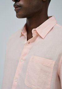 Camisa de botones de color rosa claro, hecha de lino, con un bolsillo en el pecho izquierdo. Cuello corto y textura sutil visible.
