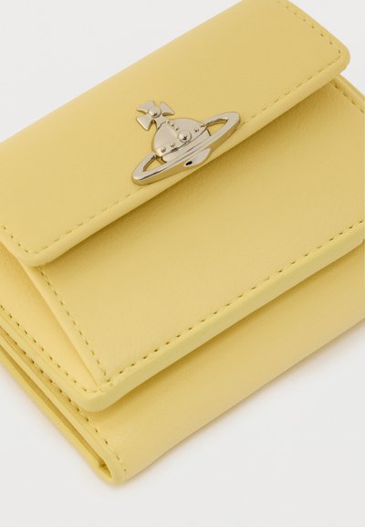 Κίτρινο δερμάτινο πορτοφόλι με διπλωτό σχέδιο, που διαθέτει μεταλλική αγκράφα σε σχήμα πλανήτη. Λείο υλικό και λεπτομέρειες με κορυφαίο ράψιμο.