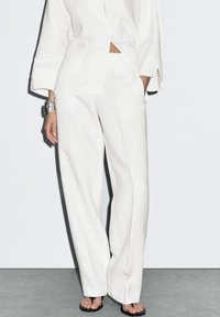 Weißes Leinenhemd und weit geschnittene Hosen mit entspanntem Sitz und Knopfdetails. Kombiniert mit schwarzen Flip-Flops und einem silbernen Armband.