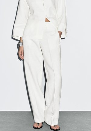 Hvid hørskjorte og bukser med vide ben, med en afslappet pasform og knapdetaljer. Sammen med sorte klipklappere og et sølvarmbånd.