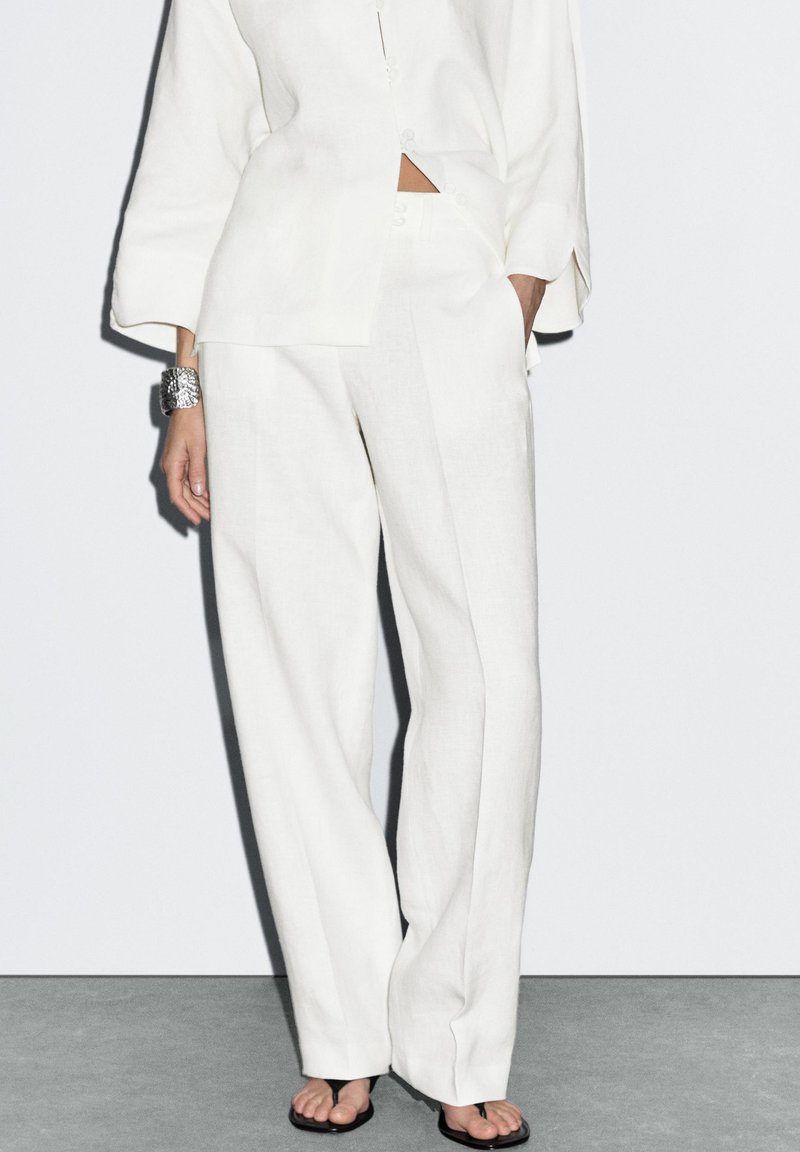 Weißes Leinenhemd und weit geschnittene Hosen mit entspanntem Sitz und Knopfdetails. Kombiniert mit schwarzen Flip-Flops und einem silbernen Armband.