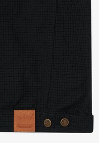 Čierna textúrovaná tkanina s prešitým vertikálnym švom, dvoma kovovými gombíkmi označenými BLKVIS a hnedou koženou nášivkou s textom značky.