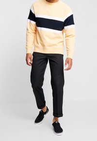 Man som bär en beige sweatshirt med svart- och vitrandigt mönster, svarta byxor med uppvikt fåll och svarta Nike-sneakers, stående på ett vitt golv.