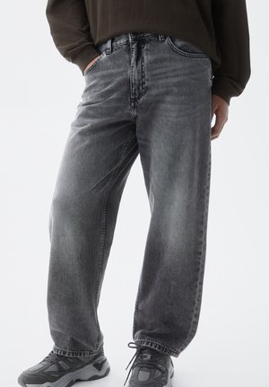 Osoba ubrana w luźne, wyblakłe czarne dżinsy, ciemną bluzę i czarno-szare sneakersy, stojąca z częściowo wsuniętymi rękami do kieszeni.