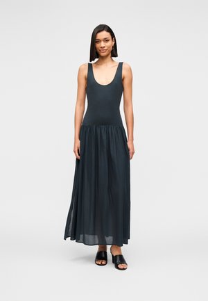 Robe maxi gris foncé sans manches avec un corsage ajusté et une jupe volumineuse et fluide. Associée à des sandales noires à enfiler. Texture lisse.