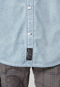 Vaalean sininen napitettu paita, jossa on musta etiketti, harmaista ja ruskeista sävyistä koostuvien ruutuhousujen päällä.