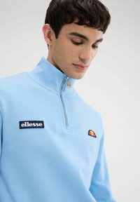 Sudadera con cierre de color azul claro, fabricada en una mezcla de algodón. Presenta un parche con el logo y un detalle bordado multicolor en el pecho.