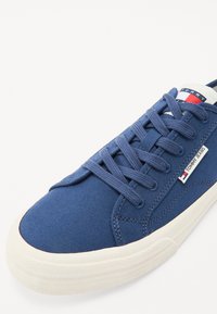 Tengerészeti vászon sneaker fehér gumi talppal, kék fűzőkkel és vörös-fehér logó cimkével a nyelv közelében. Texturált szövetfelület.