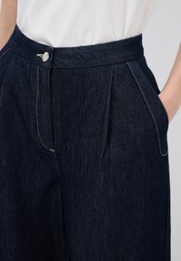 Tummat farkut, joissa on laskostettu etuosa, sivutaskut, napin kiinnitys ja näkyvää kontrastiväristä ommelta. Suunnaltaan sileä rakenne.