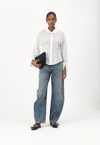 Chemise blanche à manches longues avec col, associée à un jean denim bleu ample. Pochette noire texturée et ballerines noires avec des accents en nœud.