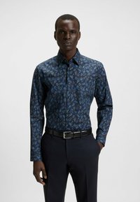 Langarm overhemd van donkerblauwe stof met een blauw en groene bladvormige print. Met knoopsluiting en aansluitend ontwerp, gedragen met donkere broek en riem.