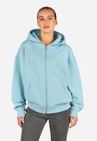 Világoskék cipzáras kapucnis pulóver, tágas elülső zsebbe, bordás mandzsettákkal és hímzett logóval a mellkason; puha pamut anyagból készült.