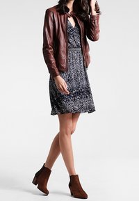 Frau in dunkelblauem Blumenkleid, brauner Lederjacke und braunen Wildleder-Stiefeletten, die mit einem Fuß nach vorne steht.