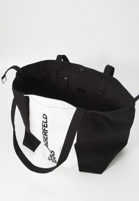 Black and white canvas tote bag with sturdy handles, featuring an interior zipper pocket and graphic text on one side. Textured fabric.