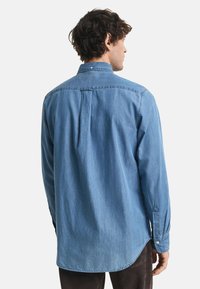 Camicia in denim azzurro chiaro con colletto classico, maniche lunghe e dettaglio a piega sul retro. Presenta polsini con bottoni e orlo curvo.