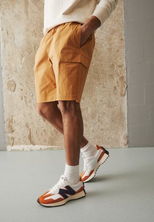 Homme portant un short cargo beige, des chaussettes blanches et des baskets blanches avec des détails orange et marine, debout contre un mur texturé.