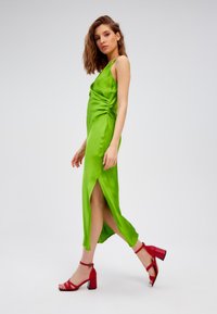 Groene satijnen jurk met een zijopening en geplooide details, gecombineerd met rode sandaaltjes met blokhak. Eenvoudig, elegant ontwerp met een gladde textuur.