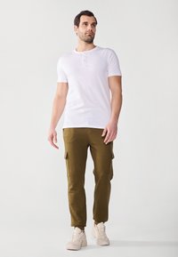 Weißes kurzärmliges Henley-Shirt mit drei Knöpfen, kombiniert mit olivgrünen Cargo-Shorts mit seitlichen Taschen und schmal zulaufenden Bündchen, getragen mit hellen Sneakers.