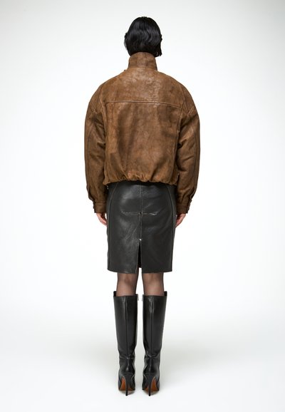 Rövid fekete hajú személy barna velúr dzsekit, fekete bőr szoknyát hátsó sliccel, átlátszó fekete harisnyát és fekete térdig érő csizmát visel.