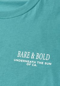 Gros plan sur un tissu bleu sarcelle montrant le texte blanc « BARE & BOLD » et « UNDERNEATH THE SUN OF CA. » imprimé sous l'encolure d'une chemise.