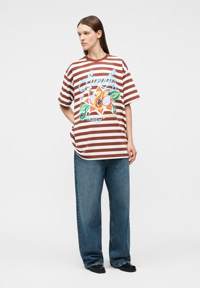 Γυναίκα που φοράει καφέ και λευκό ριγέ υπερμεγέθη μπλουζάκι με γραφικό φρούτου και μπλε κείμενο, συνδυασμένο με φαρδιά μπλε τζιν και μαύρα παπούτσια.