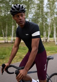 Maillot de cyclisme noir avec une bande blanche sur les bras, associé à un short bordeaux. Texture lisse, design ajusté, et vélo sous les mains.