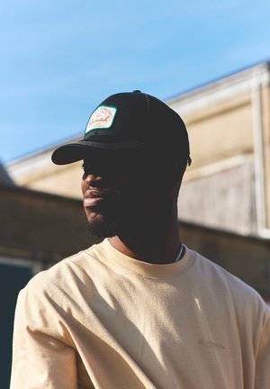 Sort baseball cap med en grøn og hvid mærkat, båret med en lys beige sweatshirt. Kasketten har en buet skygge og struktureret stof.