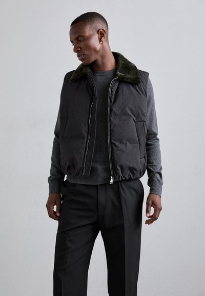 Črn napihnjen jopič s temno zelenim krznom, zadrgo in dvema stranskima žepoma, oblečen čez sivo pulover in črne hlače.