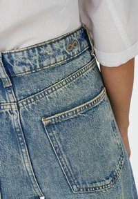 Lichtblauwe denim jeans met een hoge taille, voorzien van gele stiksels en een oneindigheidssymbool op de achterste tailleband. Twee zichtbare achterzakken.