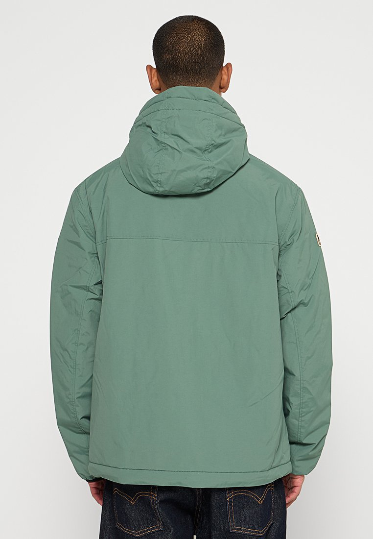 Veste isolée verte avec capuche, texture unie, poignets élastiques et détail de couture à l'arrière, portée sur un jean foncé.